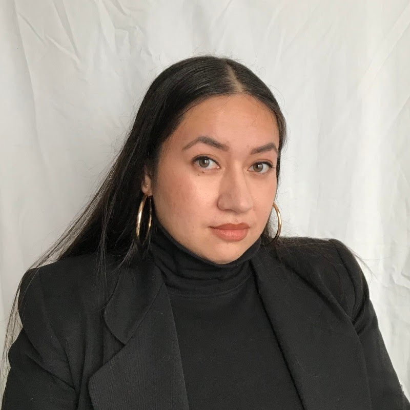 Portrait of Anna Balmilero, a woman with light brown skin and long dark hair with a part down the middle. She is looking into the camera and wearing a black blazer and black turtleneck against a white backdrop.