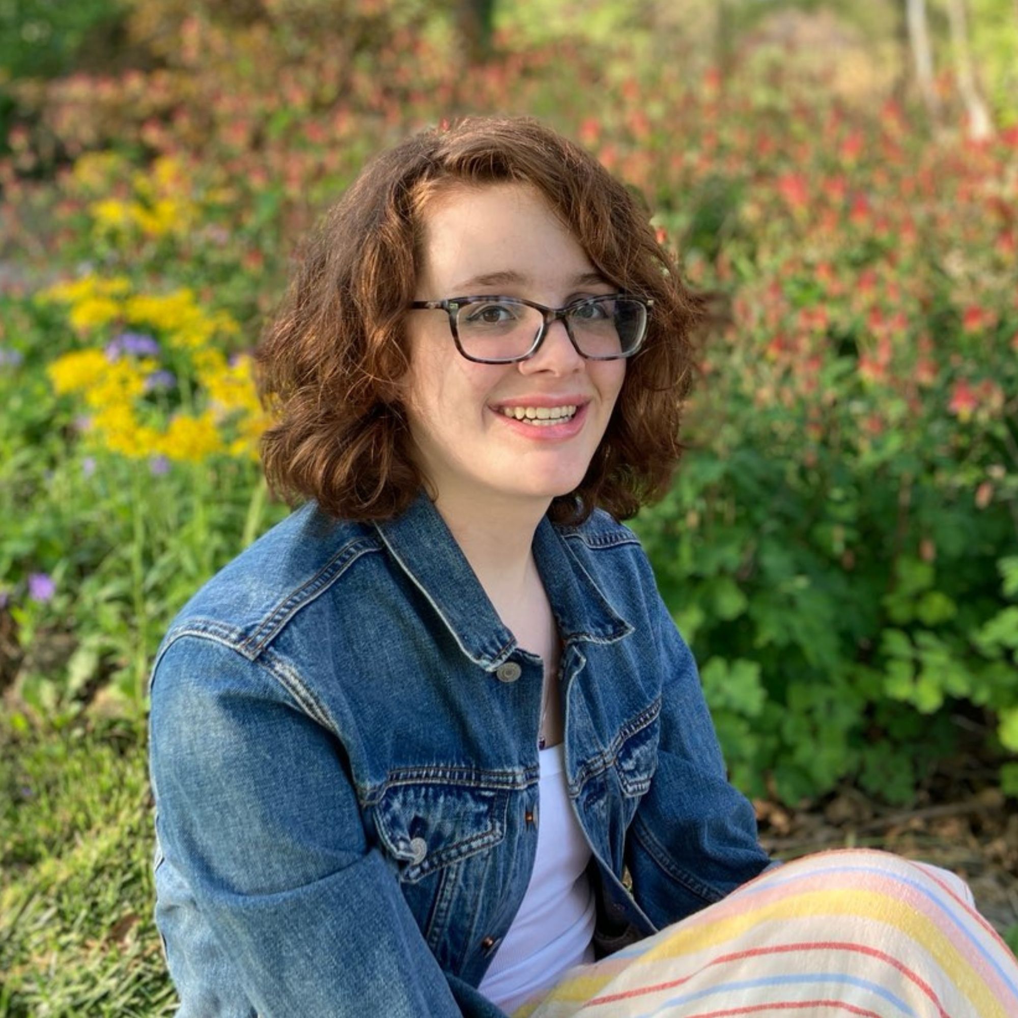 Jenna Bellamere portrait; a white woman with shoulder-length reddish brown hair, glasses, and a denim jacket sits in front of flowers, smiling