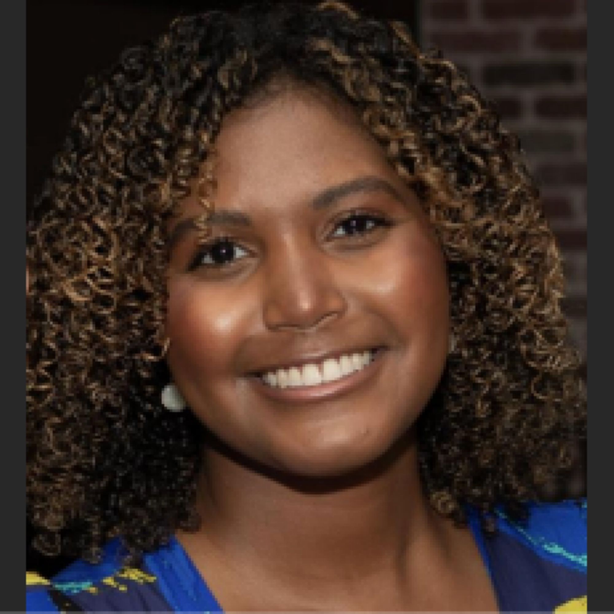 Jasmine McBride portrait, Woman of Color with medium brown skin and dark hair with tight waves and natural highlights, wearing blue, in front of brick wall